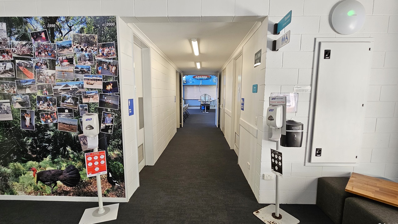 The corridor is carpeted in a dark color and lined with white walls. On the left wall is a large collage of photographs pinned close together, featuring images of people and activities. Hand sanitizing dispensers are placed on both sides. Toilet signage is placed on both sides of the wall. An open door leads into a bright, blue-coloured room at the far end of the corridor.