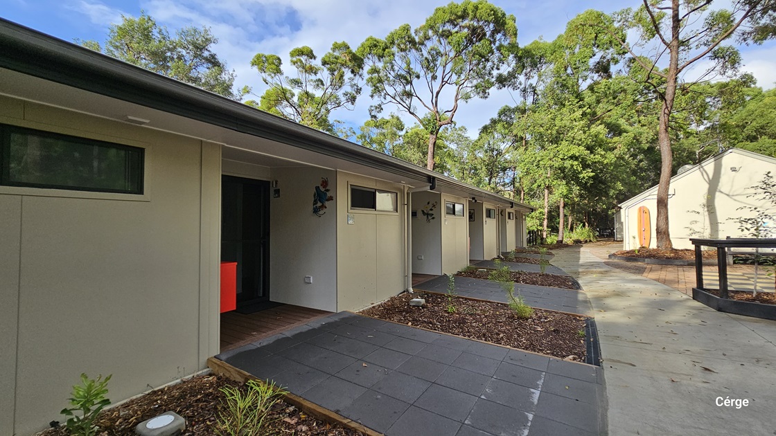On the left, there is a signpost with directions to Acacia and Banksia, next to a beige building partially obscured by trees. Two orange traffic cones are placed near a utility box. On the right, a black railing surrounds a mulched garden bed in front of a white building with a covered entrance. The path between the two areas is paved with bricks and concrete, leading into a wooded area in the background. The floor is a mix of brick and concrete.