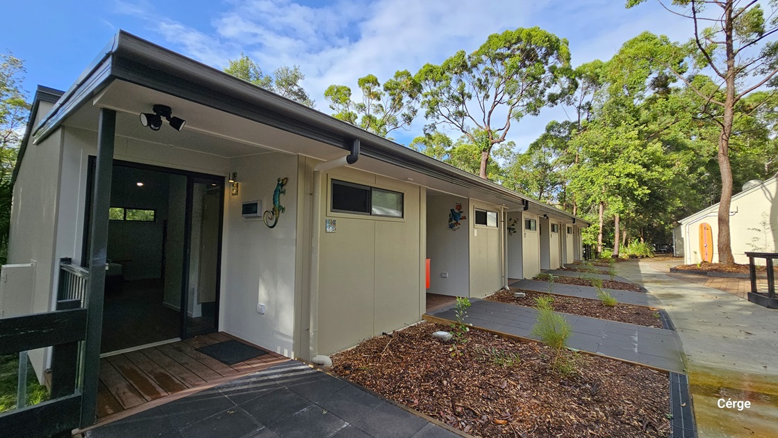 On the left, there is a row of modern, single-storey buildings with light-coloured walls and small windows. Each unit has a small porch area with wooden decking. In the middle, there is a narrow pathway with dark tiles leading along the front of the buildings. On the right, there is a paved area with a few small plants and trees. The background features tall, green trees and dense foliage.