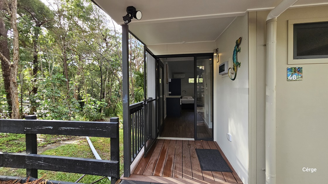 On the left, there is a black wooden railing with a view of a green, wooded area with various trees and plants. On the right, a beige wall with a colourful gecko decoration and a room number sign "48". In the middle, a glass sliding door leads to an interior space with a visible bed. The foreground features a wooden deck floor with a black doormat. The ceiling is white and has a light fixture.