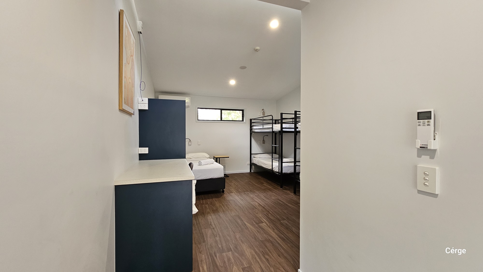 A room with a timber floor and white ceiling with recessed lighting. On the left, there is a blue and white cabinet with a framed photo above it. In the middle, a single bed with white bedding is positioned against the wall. On the right, there are two black metal bunk beds. At the end, there's a rectangular window with a view of greenery. The foreground includes a white wall with a thermostat and light switch.