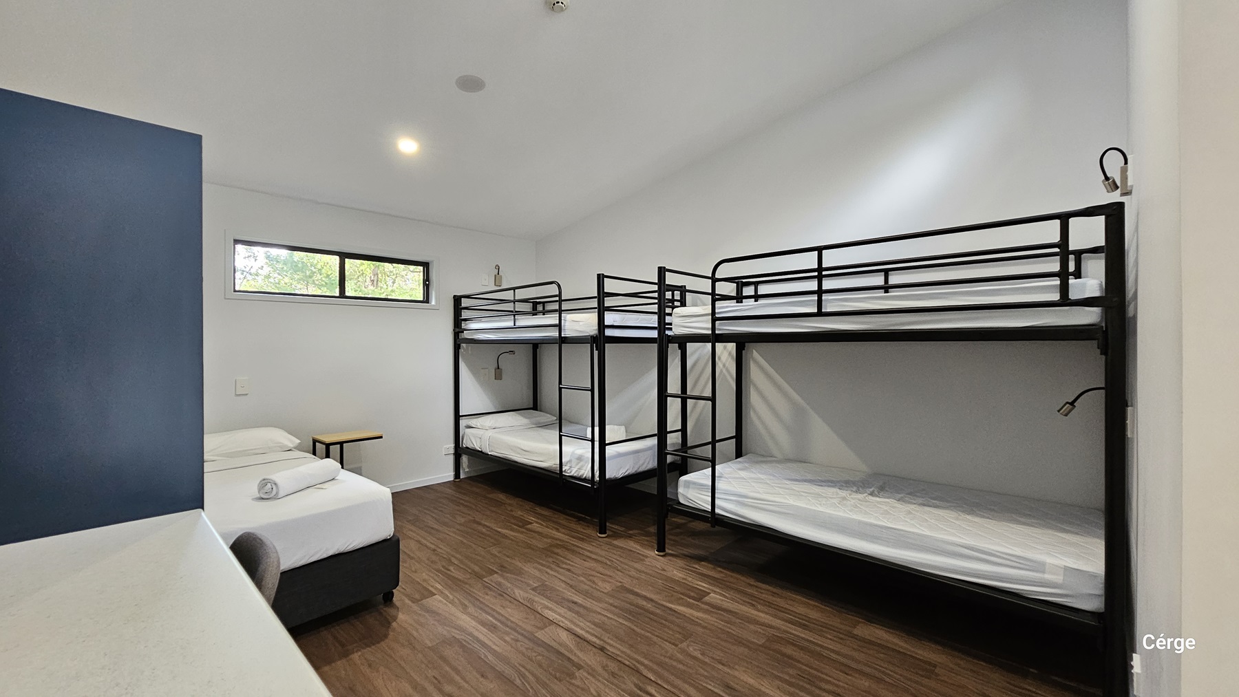 On the left, there is a single bed with white sheets and a pillow, next to a small wooden table. On the right, there are two sets of black metal bunk beds with white mattresses. In the middle, there is a small window with a view of trees. The floor is made of dark timber, and the ceiling is white with a recessed light. The walls are white, except for a dark blue section on the left.
