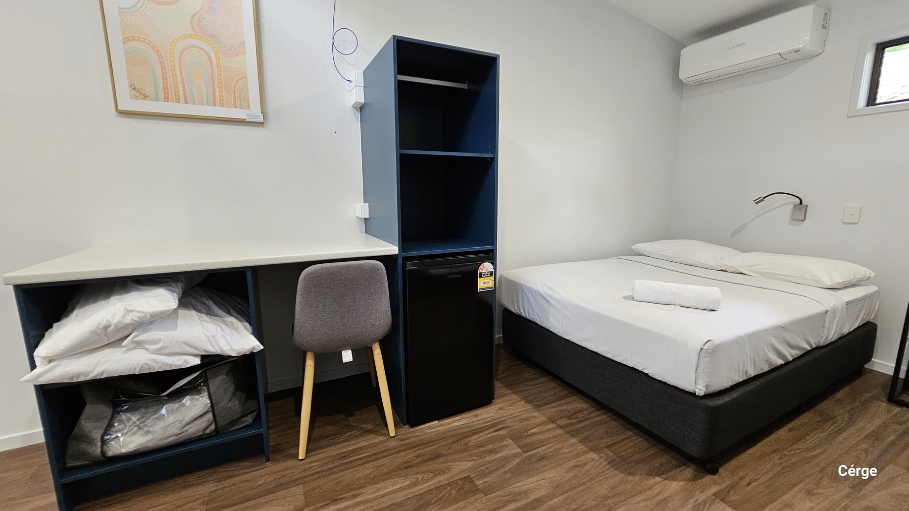 On the left, there is a white desk with a grey chair in front of it. Under the desk, there are shelves containing pillows and a blanket. Next to the desk is a small black refrigerator. In the middle, there is a blue shelving unit. On the right, there is a bed with white sheets and a towel on top. Above the bed, there is an air conditioner mounted on the wall. The floor is timber, and the ceiling is white.