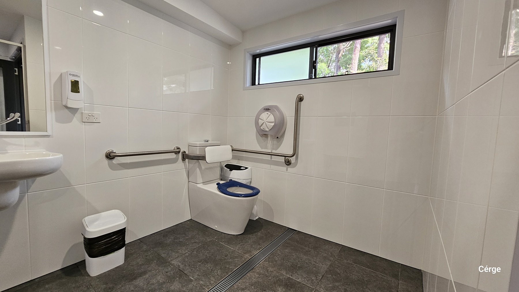 On the left, there is a white sink with a soap dispenser mounted on the wall above it. Below the sink, a small white bin with a black liner is placed. In the middle, a white toilet with a blue seat is installed, accompanied by stainless steel grab bars on both sides. On the right, a large toilet paper dispenser is mounted on the wall. The floor is covered with dark grey tiles, and the walls are lined with white tiles. A rectangular window is positioned high on the wall.