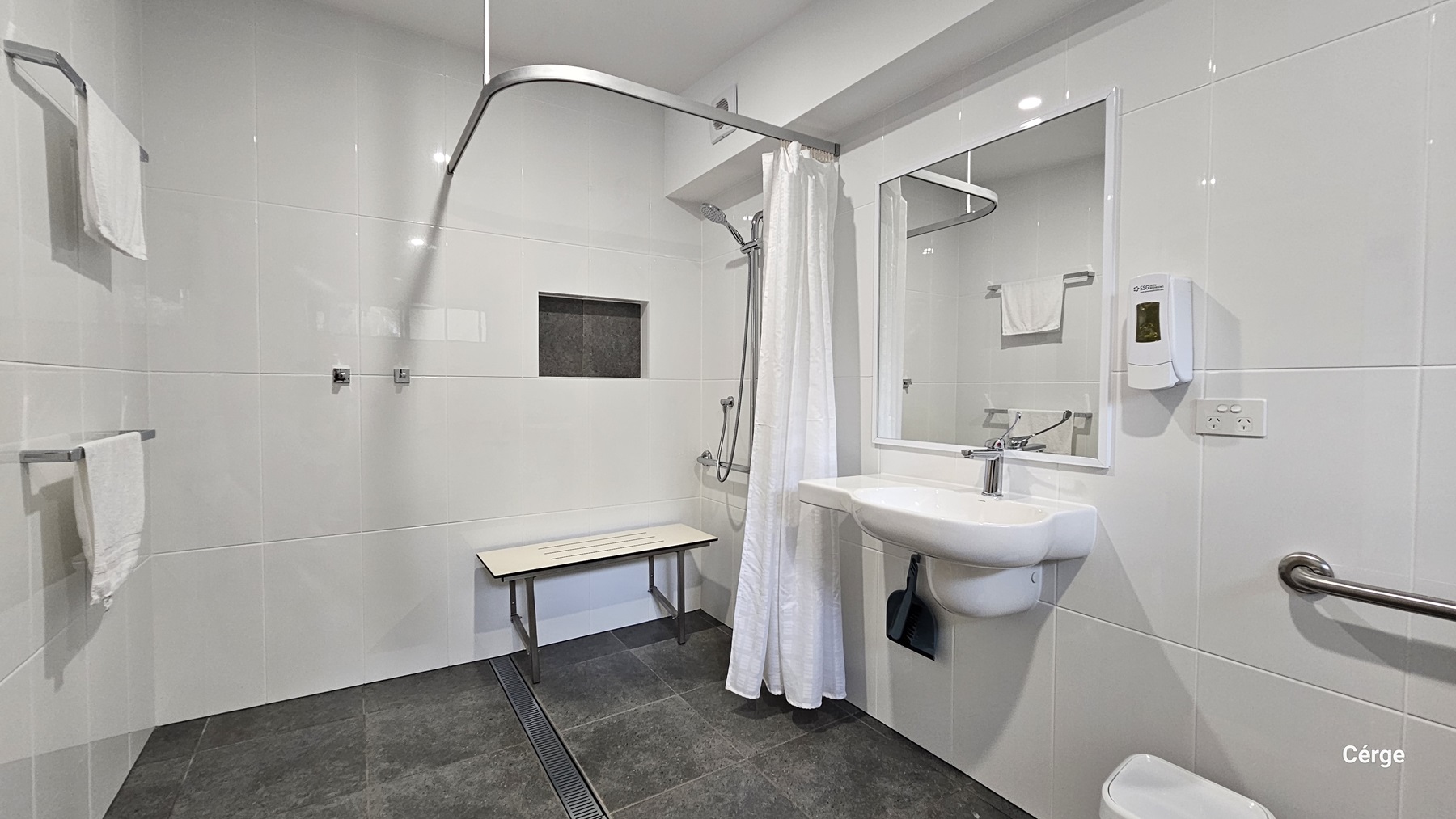A bathroom with white tiled walls and a dark grey tiled floor. On the left, there are two towel racks with white towels. In the middle, a shower area with a white curtain, a wall-mounted showerhead, and a foldable bench. On the right, a white sink with a mirror above it, a soap dispenser, and a grab bar. The ceiling is white with recessed lighting.