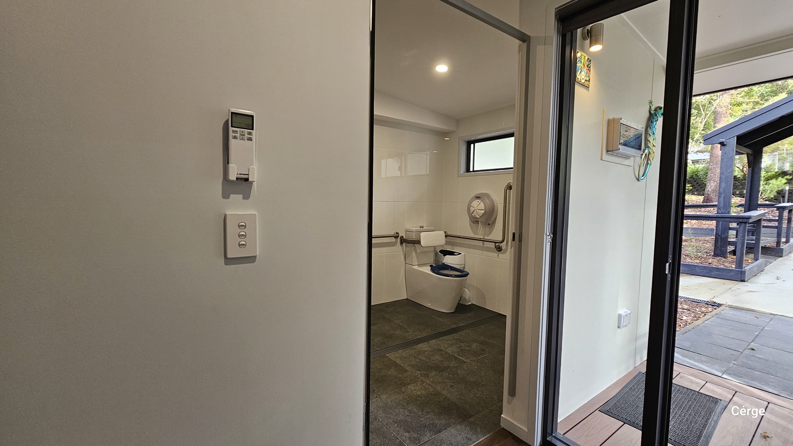 On the left is a white wall with a thermostat and a light switch. In the middle, is a bathroom with a toilet, grab bars, and a small window above. The floor is dark grey tiles. On the right, a glass door leads outside to a patio area with a black railing and a pathway. The ceiling is white with recessed lighting.