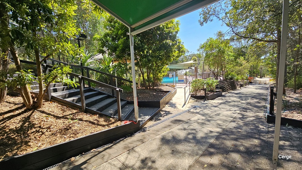 On the right is a staircase with handrails. Trees and ornamental plants are positioned on the right. A green shade cloth covers a section of the garden. In the middle is a concrete ramp with handrails leading to the pool. To the right, the road leads to the foreground, bordered by trees.