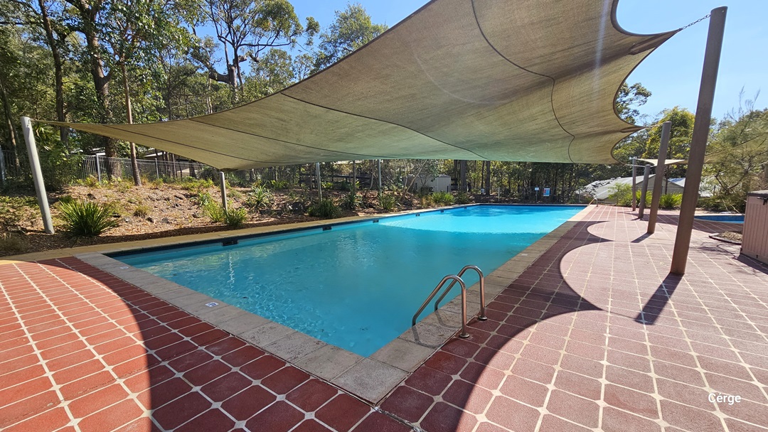 The 25m pool is equipped with handrails and a red tile pool deck. A shade cloth covers a section of the pool. Trees and shrubs surround the area. On the far right is a smaller pool.