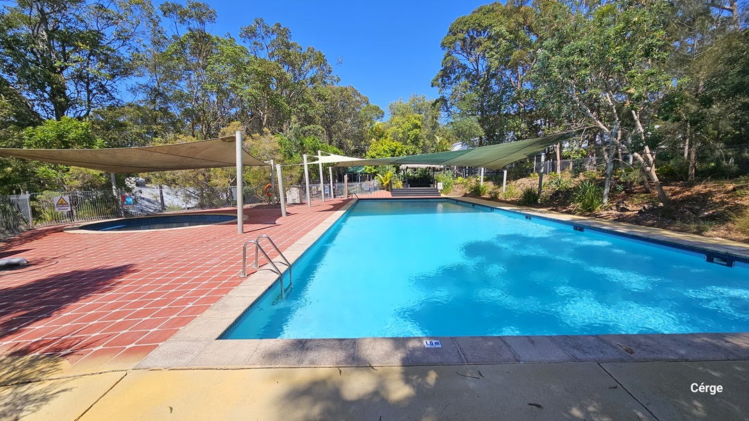 The 25m pool has handrails and a red tile pool deck. A shade cloth covers part of it. On the left is a round pool with shade cloth. Trees and shrubs surround the area. On the far right is a smaller pool. The pool area is secured by a metal fence with safety reminders affixed.