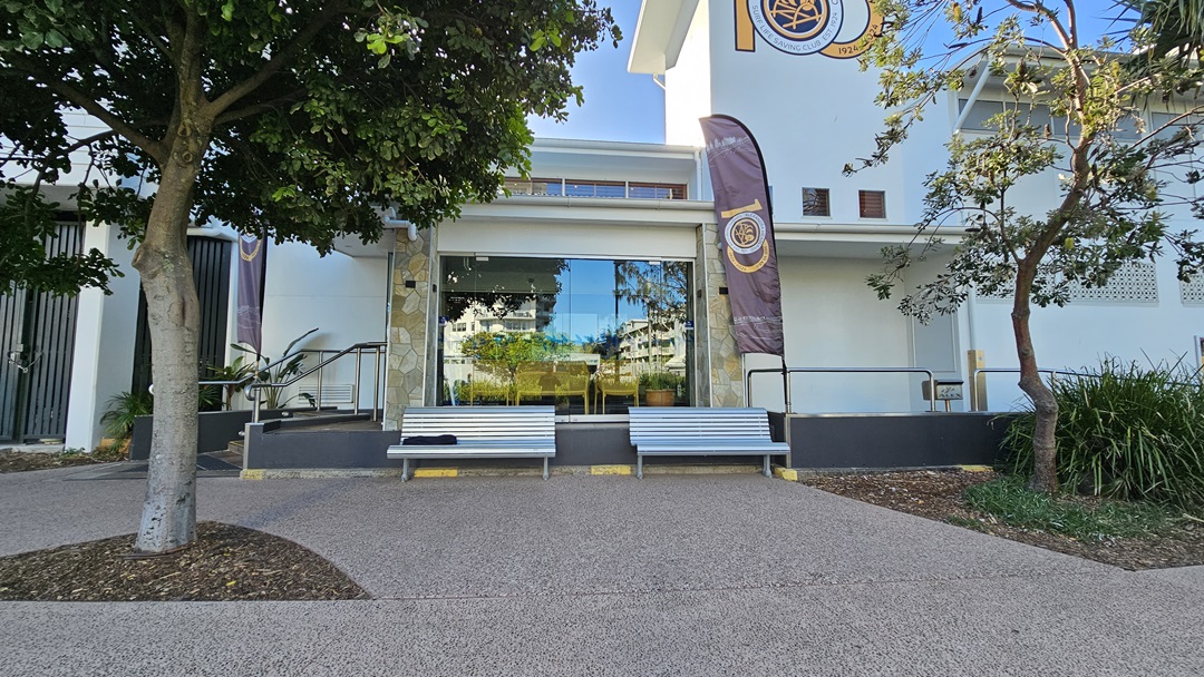 On the left, there is a tree with a thick trunk and green foliage. To the right, another tree with sparse leaves stands near a building. In the middle, a large glass window reflects the surrounding area, flanked by two grey benches with yellow accents. The background features a white building with a purple and gold flag. The floor is a textured, light brown pavement.