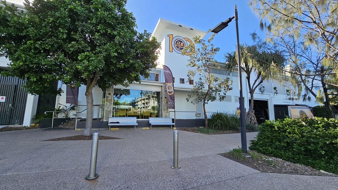On the left, there is a large green tree next to a building with a glass entrance and a ramp. In the middle, two benches are placed in front of the entrance, flanked by two vertical banners. On the right, there is another tree with sparse foliage and a bushy area. The background features a white building with multiple windows and a sign. The floor is paved with a light brown, textured surface, and there is no visible ceiling.
