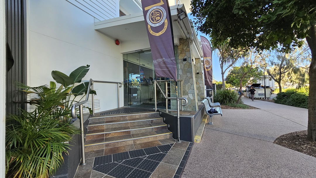 On the left, there are potted plants next to a building entrance with glass doors and a metal handrail. The middle features stone steps leading up to the entrance, flanked by two vertical banners. On the right, there is a bench and a tree with dense foliage. The background includes a pathway and more greenery. The floor is a mix of stone tiles and textured paving.