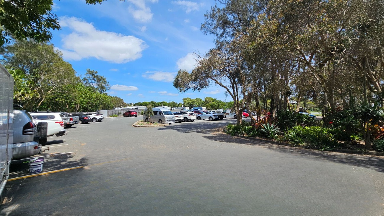 On the left, parked cars are lined along a paved parking area surrounded by green leafy trees. On the right, trees with thin trunks and thick foliage partially cover landscaped garden beds with various plants. In the middle, more parked cars, including a van, are scattered across the open paved surface. The background features more parked vehicles, fencing, and dense trees. The floor is an asphalt surface with visible wear and faint shadow patterns.