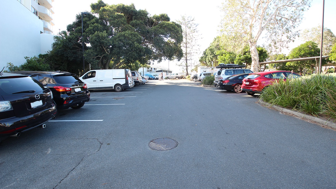 A tarmac parking area with marked spaces runs into the background. On the left, parked black cars and a white van are lined along the edge beside a tall white building and dense trees. On the right, a row of cars, including a red vehicle, is next to plants and trees. In the middle, the open tarmac features a circular manhole cover. The background includes additional vehicles, trees, and a white building visible in the distance.