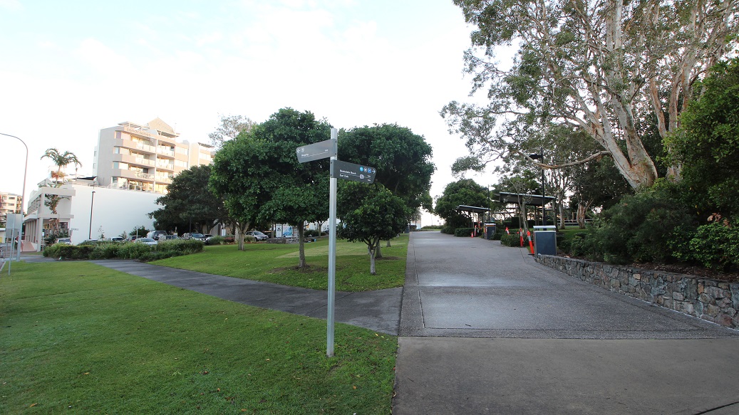 A paved path runs from the foreground into the middle, bordered by grass on the left and a low stone wall with trees on the right. On the left, a modern apartment building and parked cars are partly visible near clusters of trees. On the right, large trees with overhanging branches and shaded seating areas with structures appear alongside a bin. In the background, the path continues, flanked by trees and landscaping. The floor is a mix of grass, pavement, and concrete.