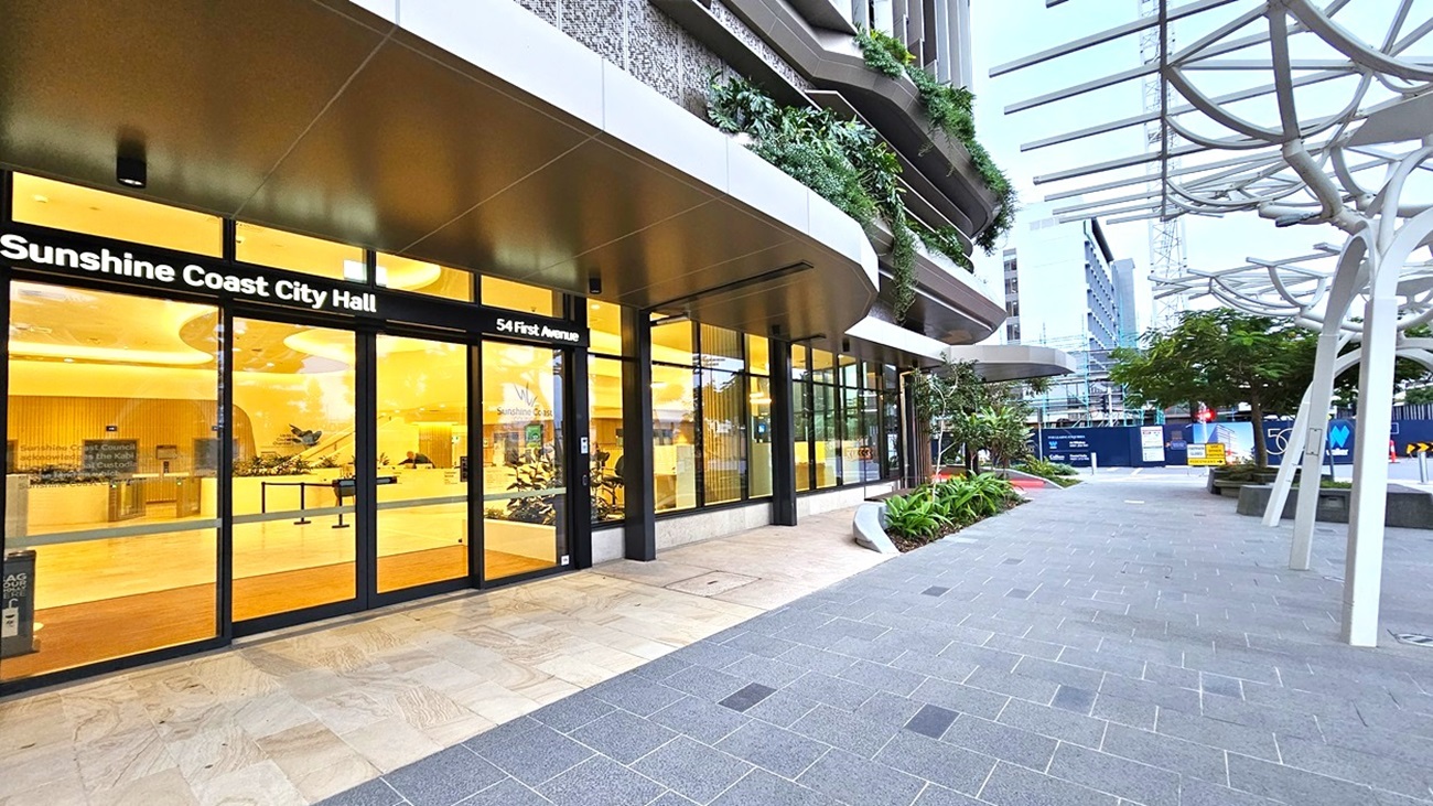 On the left, there is the entrance to Sunshine Coast City Hall with large glass doors and a sign above. The interior is brightly lit with yellow lighting. On the right, there is a walkway with grey tiles, some plants, and modern metal structures providing partial cover. In the middle, a mix of greenery and urban elements. The floor is a combination of light-coloured tiles near the entrance and darker tiles on the walkway.