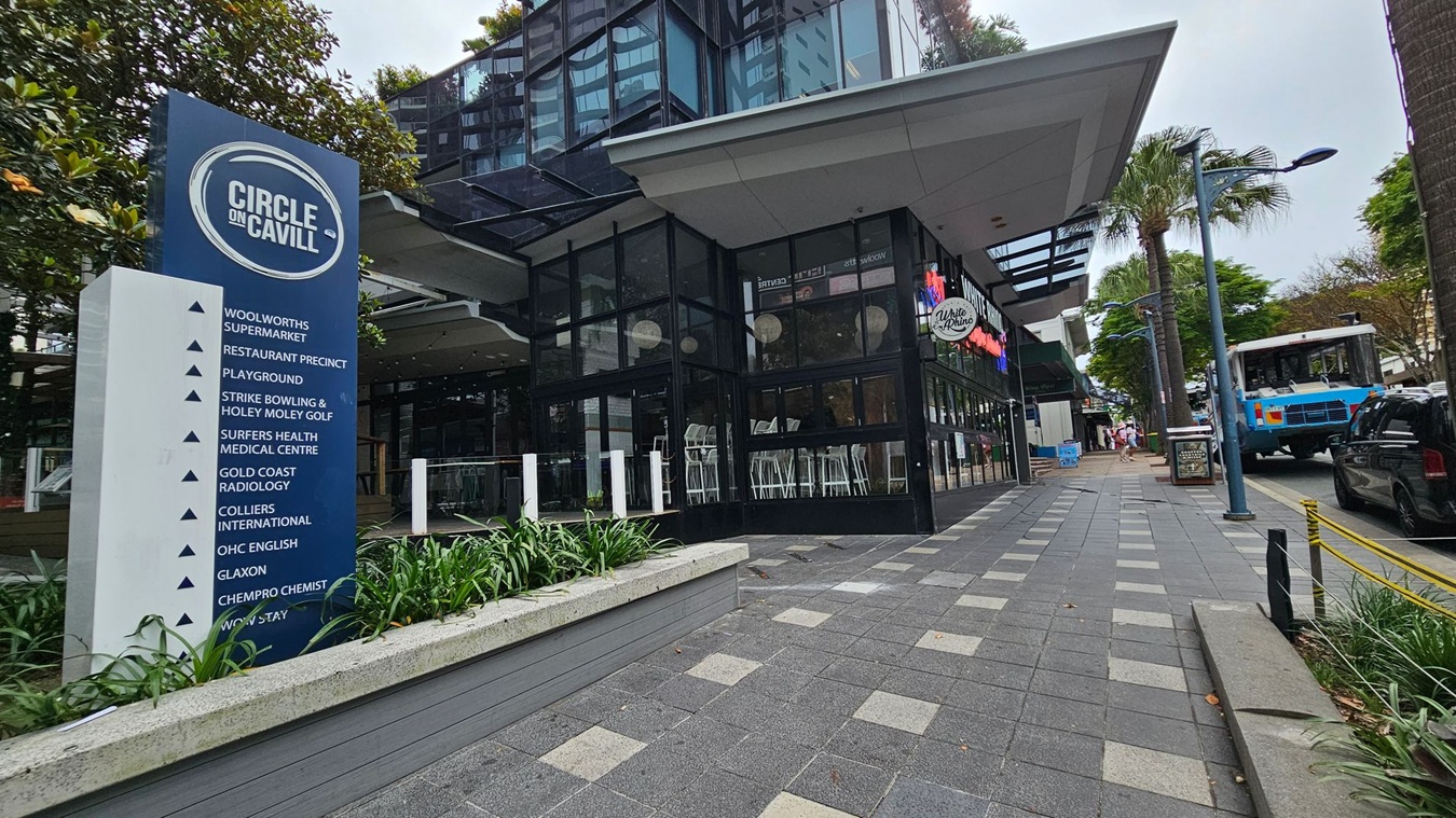 On the left, a large blue sign lists various establishments at "Circle on Cavill," including a supermarket and medical centre. In the middle, a modern building with glass walls houses a venue with high stools visible inside. On the right, a paved walkway lined with palm trees and a blue bus is parked on the street. The pavement is grey with lighter square patterns, and there are green plants in a raised bed near the sign.