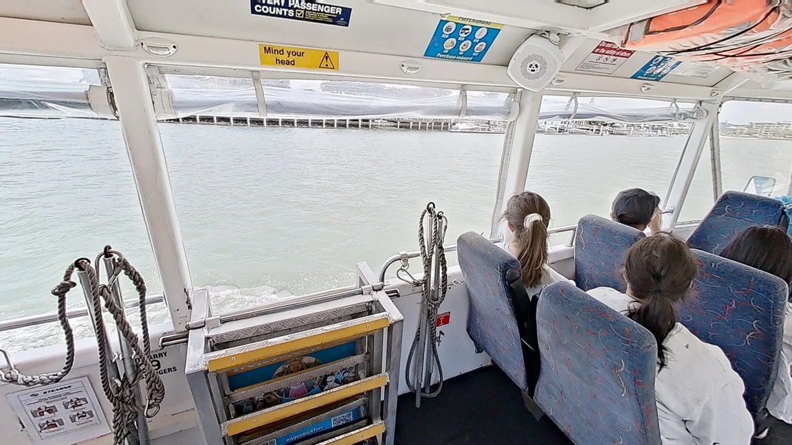 View of the Aquaduck in the water focusing on the folded 5-tiered stainless steel stair with yellow lines and rope handrails of the portion. The passengers are seated on blue seat padding with yellow, red, and light blue line patterns. The boat has a white roof with a warning sign that reads, “Mind your head,” and other safety signs and instructions on the walls. The boat has large windows that offer a view of the water and the shore.