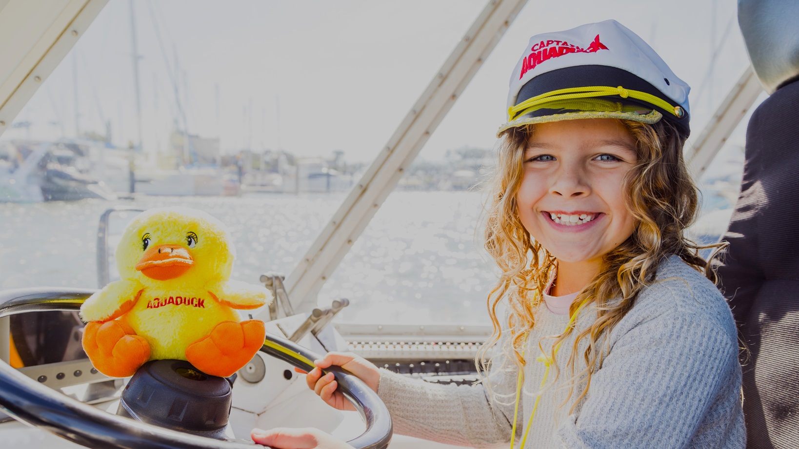 A smiling girl with blonde hair is wearing a captain's hat with white, black, pink color and yellow rope. A yellow stuffed duck with black AQUADUCK lettering in the middle of its chest is placed on top of the black steering wheel. The girl is wearing a blue netted sweater and is sitting on a grey seat with a black cover.