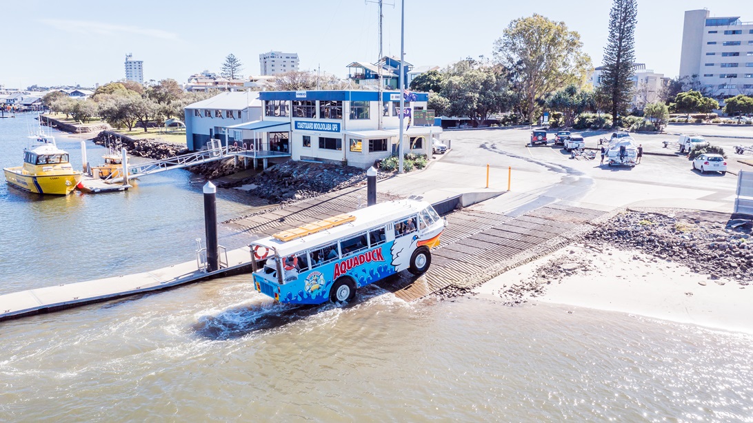 The Aquaduck, leaving the water and ascending to a steep boat ramp that has horizontal lines. There is a building to the left with white and blue color schemes. It has a bridge with metal handrails that connects to a boat dock. A yellow boat can be seen on the dock. The park with trees, the parking area, and the pathway leading to the main facility can be identified from this image.