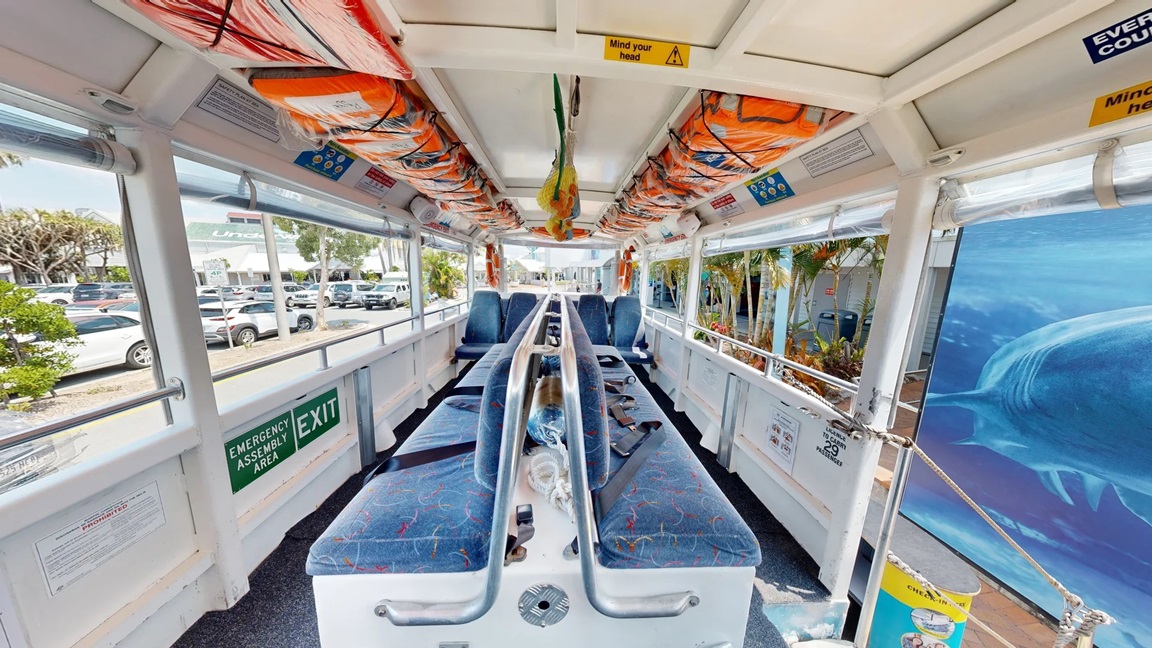The middle of the Aquaduck is a long bench-type seat with a back support that has blue seat padding with yellow, red, and light blue line patterns. To the back is another row of 4 blue seats with yellow, red, and light blue line patterns, which are separated by a center aisle. Above and below the windows are varying colors and sizes of signs for rules and safety precautions. On the ceiling are two lanes of orange life vests inside plastic secured by black straps forming an X.