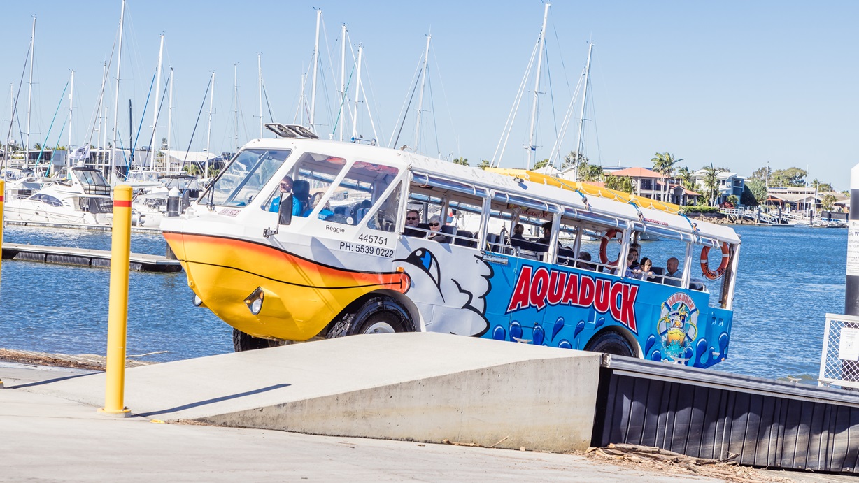 Aquaduck ascending from the steep boat ramp. Image also shows the yellow bollard at the start of the short ramp of the middle bridge. From this image, the short ramp is made of concrete, while the bridge is supported by a grey frame on the side and a grey structure underground. The back of the image shows the residential area and a marina filled with sailboats.
