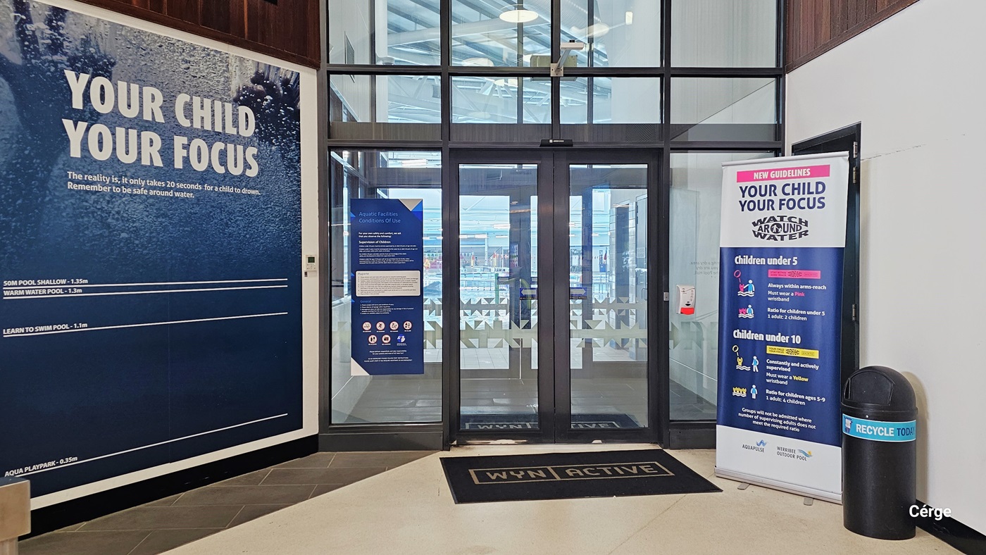 The Aquatic Centre entrance features two sets of automatic glass doors adorned with snowflake prints. A blue poster is affixed to the left section of the glass. A black doormat with "WYN | Active" is placed at the entrance. On the left, a large blue poster covers a significant portion of the wall. To the right, a banner displays child safety instructions, accompanied by a black rubbish bin. This view offers a glimpse of the indoor pool.