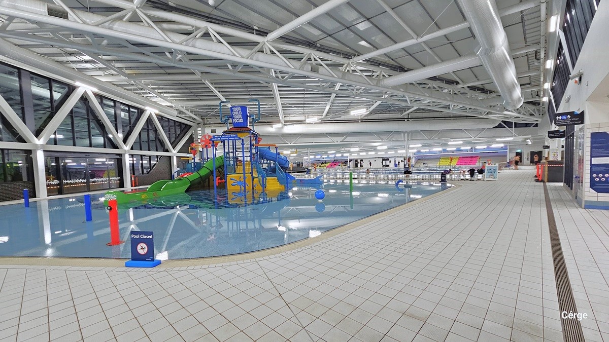 Here there is a shallow beach entry pool with two enclosed tube slides. The pool is equipped with interactive water cannons and a blue tipping bucket positioned overhead. The prominent white ceiling shows white pipes and tubes. There's a freestanding blue warning sign that reads "Pool Closed." The floor is tiled in white. On the left, a glass wall features a large steel "W" design suspended from the ceiling. To the right, banners are on display, providing instructions and directions.