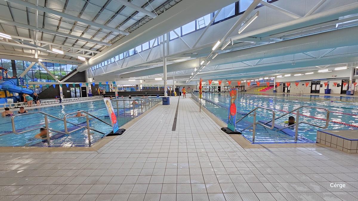 There are two heated pools separated by a white-tiled pool deck. Both pools have stainless steel entrance ramps, each adorned with two freestanding safety banners before the entrance ramps. On the left, there's a glimpse of the giant winding water slide, while on the right, multiple doors are present, alongside a tiered grandstand featuring yellow and red hues. The floor is covered in white tiles, and the ceiling is entirely white, with large pipes and visible metal beams supporting the roof.