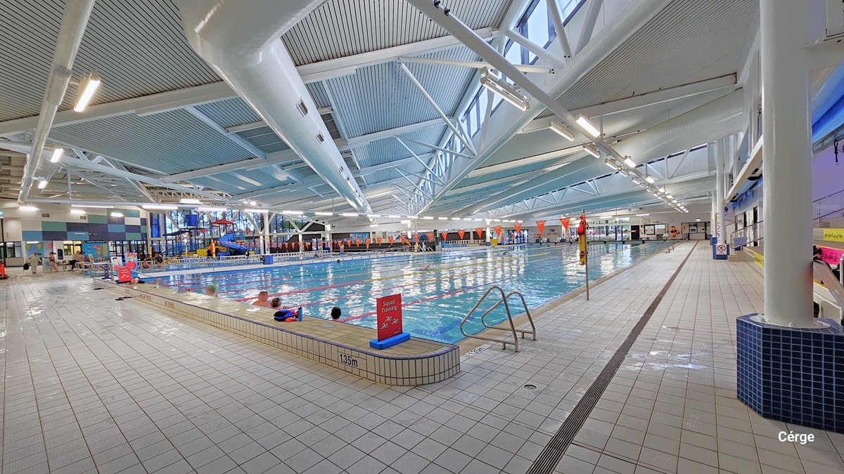 An alternate perspective of the heated pool reveals a stainless steel entrance ramp, accompanied by a freestanding orange safety banner positioned before the entrance. On the far left, a segment of the colossal winding water slide and the vibrant ceiling are visible. Orange triangular banners hang across the pool. On the right, there's a path leading to the opposite side of the pool. The floor and ceiling are both white, with large pipes and visible metal beams supporting the roof.