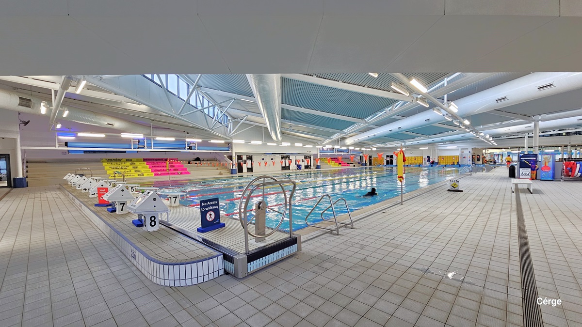 Another view of the heated pool displays a stainless steel entrance ramp and a platform featuring a stainless steel steering wheel, a freestanding safety sign, 8 stairs, and diving boards at the center. On the far left, there's a tiered platform with pink and yellow accents. To the right, additional safety signs and banners are visible, along with a chair placed in the middle. The floor and ceiling are both white, with large pipes and visible metal beams supporting the corrugated design roof.