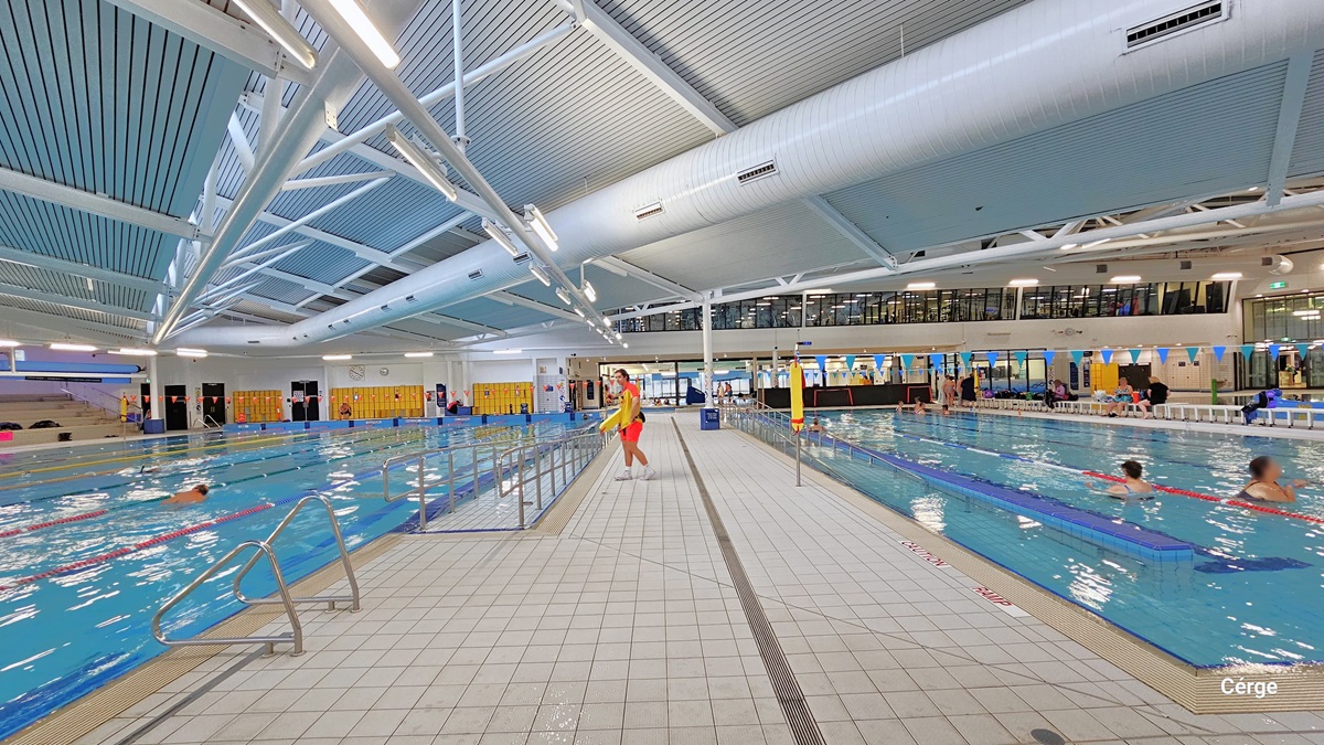 There are two heated pools, each separated by a white-tiled pool deck. The left pool features stainless steel entrance ramps, each accompanied by two freestanding safety banners and handrails. Floating ropes in various colors serve as guides. Colorful blue triangle banners hang across the pool on the right. The floor is tiled in white, and the ceiling is entirely white, with large pipes and visible metal beams supporting the roof. A few individuals can be seen swimming.