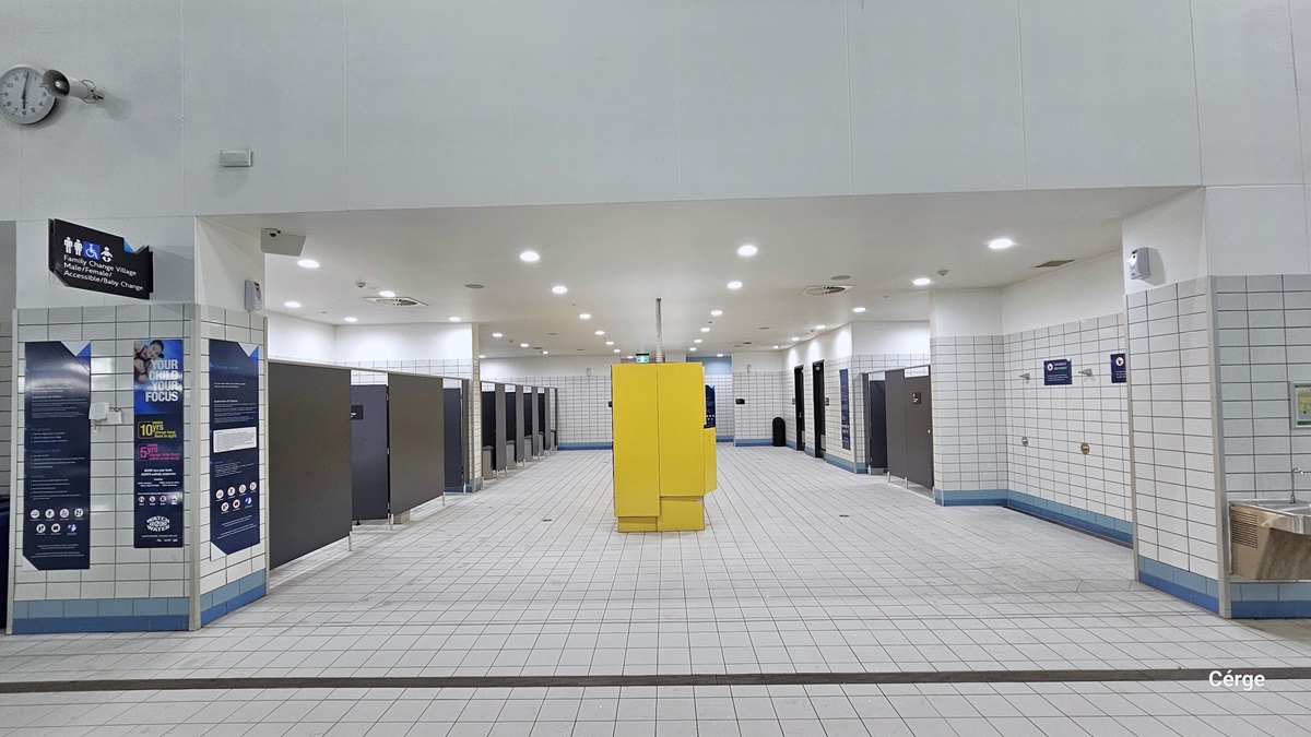 Accessing the family change room, which is a spacious area with rows of bathroom stalls enclosed by black walls and doors. In the upper left corner, there's a sign indicating gender-specific, accessible to disabled, and a baby change area, accompanied by a clock and a speaker. In the middle stands a yellow cabinet. On the right, there is another row of bathroom stalls, including a shower area. The entire bathroom is adorned with white tiles, and the ceiling is entirely white.