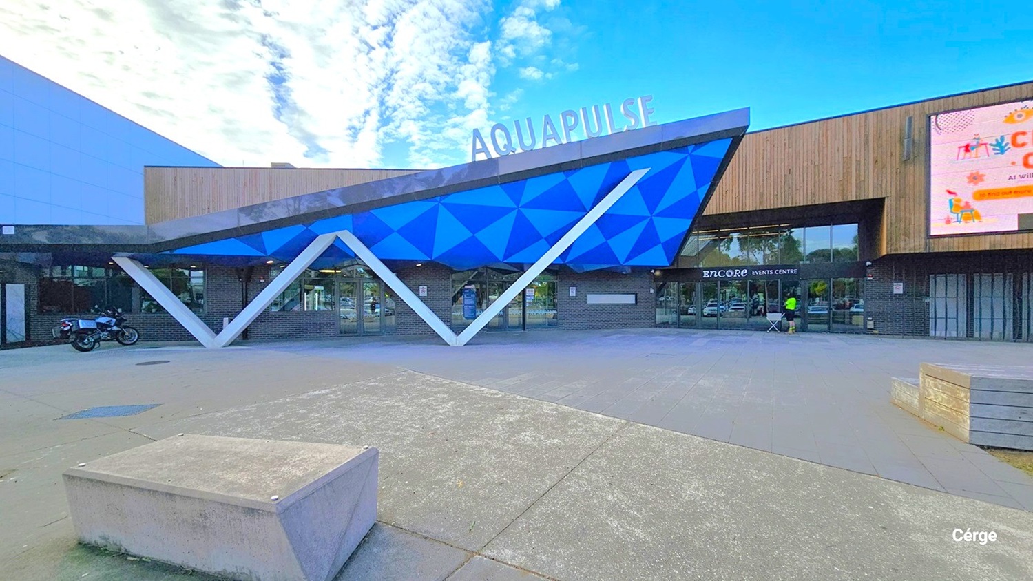 AquaPulse's main entrance has a distinctive design with the letter "W" seemingly supporting a triangular ceiling in shades of blue and green. The name "AquaPulse" is prominently displayed on the rooftop. On the right, a large digital screen is visible. In front, there's a spacious area with a raised concrete structure in the center. The building's exterior features a combination of grey and brown wooden design. A motorcycle is parked to the left.