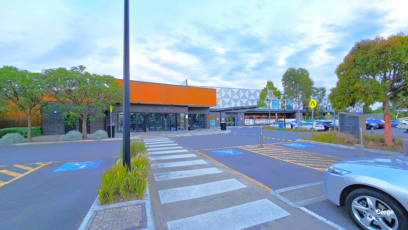 This view reveals designated open car park spaces reserved for disabled individuals, demarcated by yellow road markings and bollards. The car park area is divided by a pedestrian pathway and a plant box, featuring a tall black pole and ornamental plants. In the distance, road signs become visible, along with the step-free entrance. A silver car is currently parked in one of the reserved spaces. Further ahead, you'll see the opposite side of the parking area occupied by various cars.