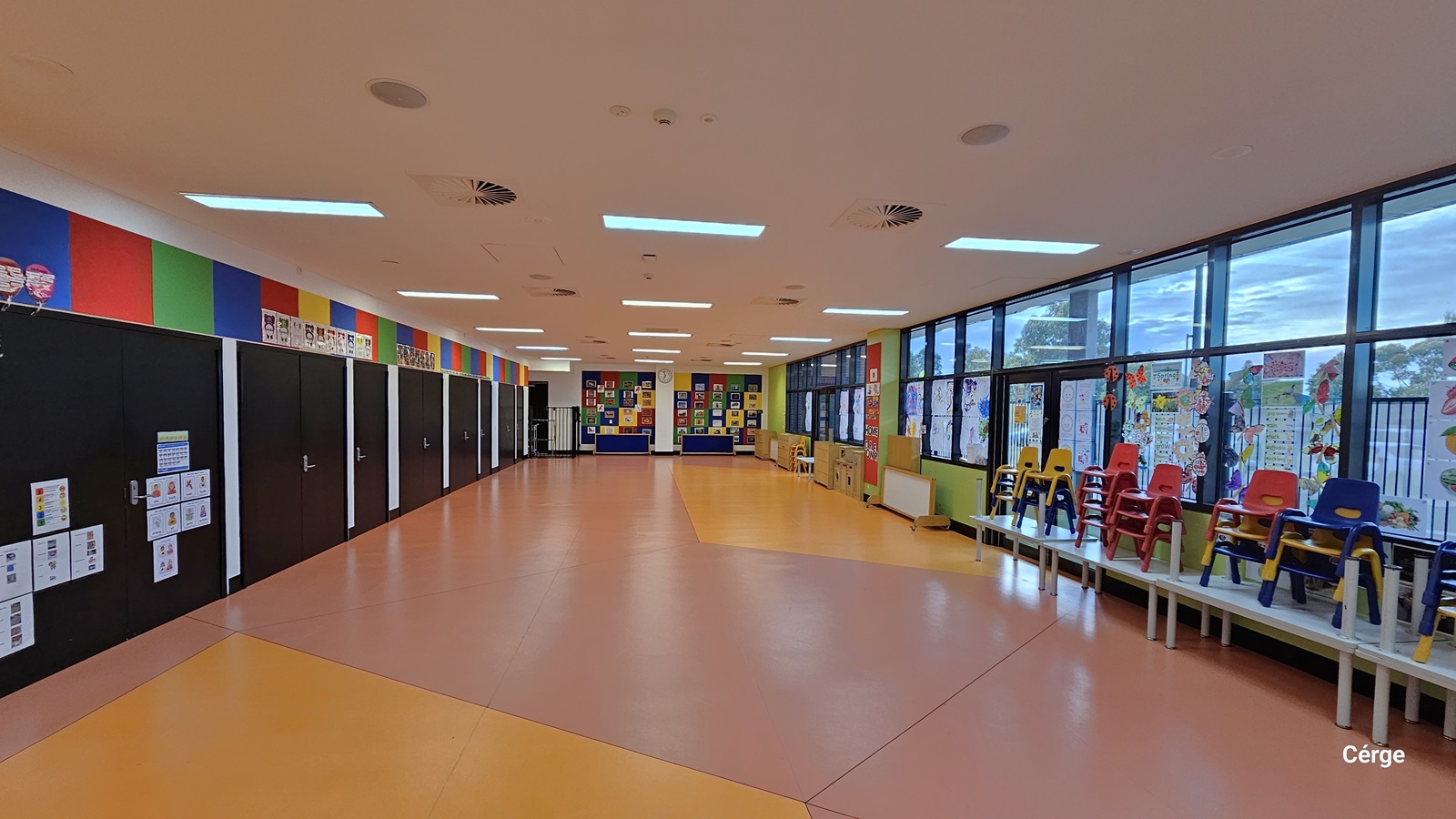 This is a large space featuring white plastic tables on the right, with a variety of stacked plastic chairs in different colors. Additional wooden tables, portable whiteboards, and cabinets are also in place. The middle wall is adorned with vibrant images and photographs, while the left wall showcases black-painted wooden cabinets with colorful posters. This well-lit area boasts a rubber floor designed with yellow and peach tiles. Large open windows offer views of the outside.