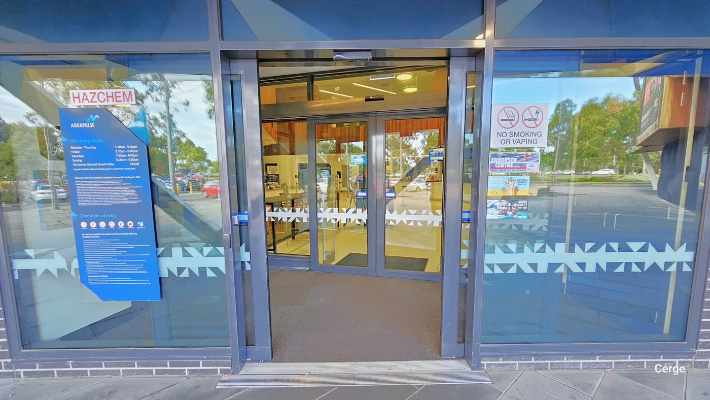 AquaPulse entrance is through two sets of automatic glass sliding doors fixed within silver railings. Stickers and banners on both doors display reminders and instructions, including a "No smoking or vaping" sign. The glass doors feature snowflake design markings. Within sight is a section of the interior cafe. whose interior is well-illuminated.