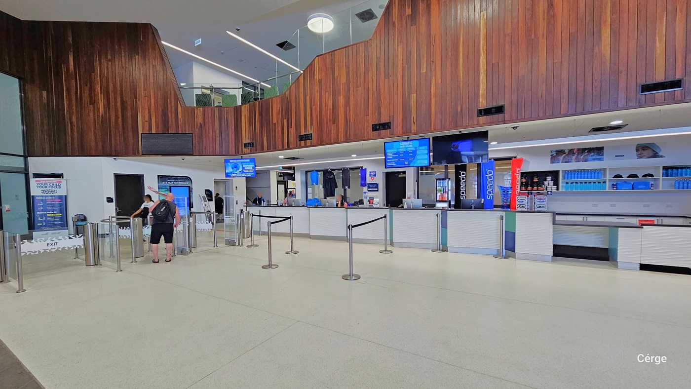 Upon entry, the reception area is straight ahead, featuring queue barriers, digital information screens, and merchandise for sale. The entrance includes a turnstile where two individuals are seen entering. The space is well-lit with a white ceiling and floor tiles. The wall showcases an intricate brown wooden design. You can also see the second-floor area from this perspective.