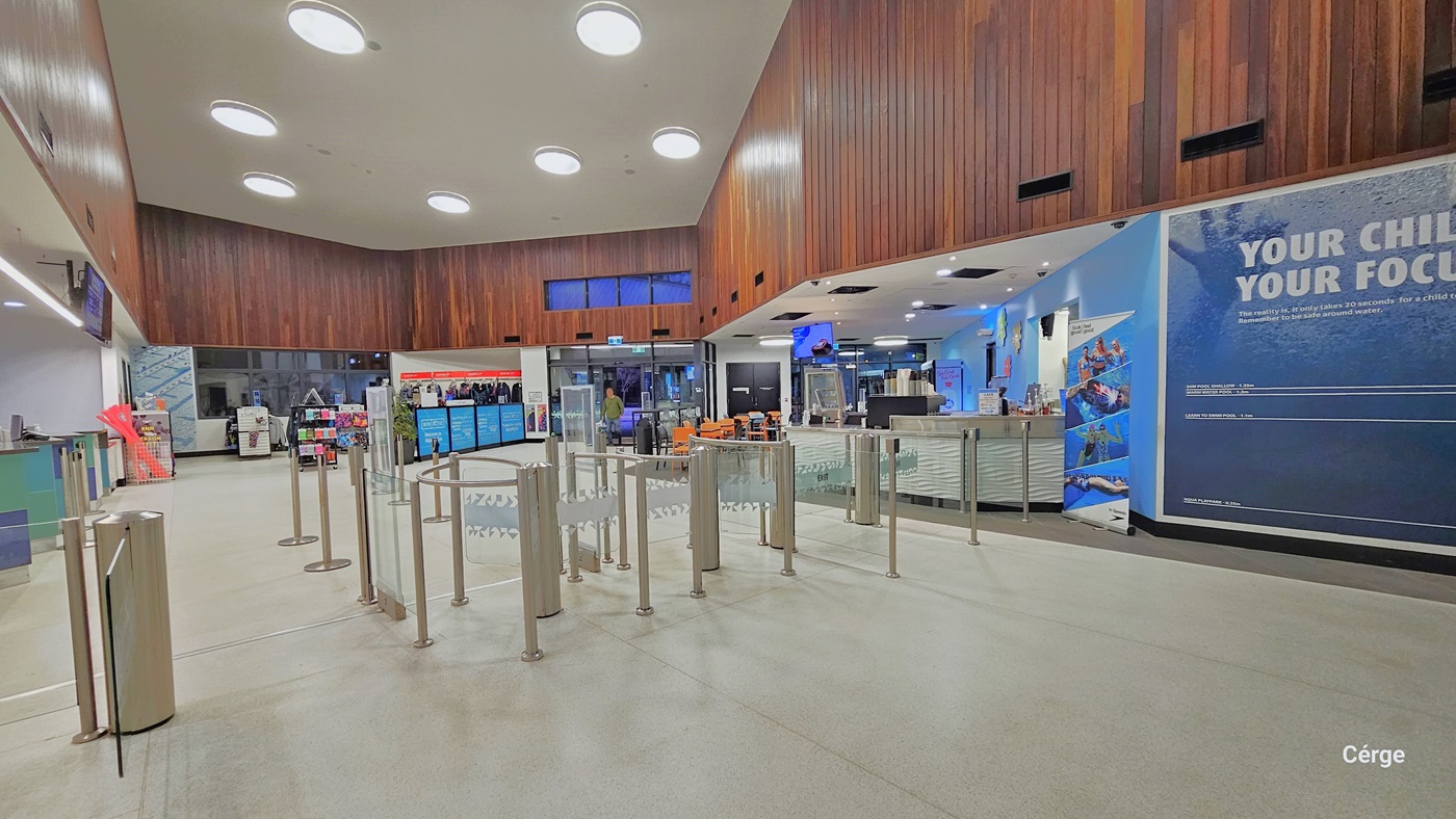 Upon entry, the reception area features queue barriers, digital information screens, and merchandise for sale. The entrance has a turnstile with two people entering. Well-illuminated space with a white ceiling, floor tiles, and an intricately designed brown wooden finish on the wall. Further ahead, a retail area offers swimming costumes, goggles, and swim accessories.