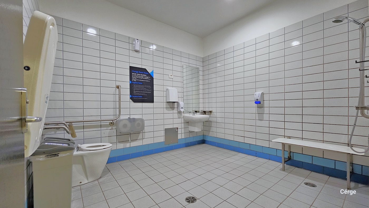 Inside the Accessible Bathroom, to the left, there is a folded changing table mounted on the wall, two rubbish bins, and the ambulant toilet, complete with grab bars. In the corner, there's a wash basin with a tissue dispenser, hand dryer, and liquid soap dispenser. Additionally, there is a hand dryer on the wall. On the right, there's a shower with a long white bench beside it. The bathroom is well-lit, with white floors, walls, and ceiling.