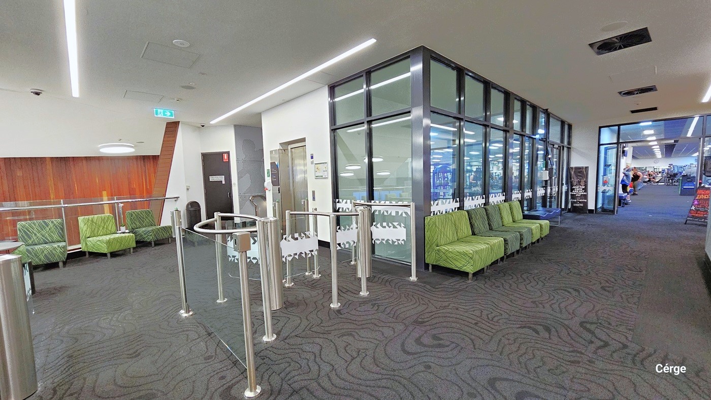 Entry to the AquaPulse gym is through turnstiles at the entrance. A few cushioned green chairs are lined up against the glass wall. From this viewpoint, there is a section of the spacious gym with colorful banners on display. The entire area has grey carpeting with a jigsaw puzzle design. Long fluorescent lights are mounted on the ceiling, along with a row of centralized air conditioning systems.