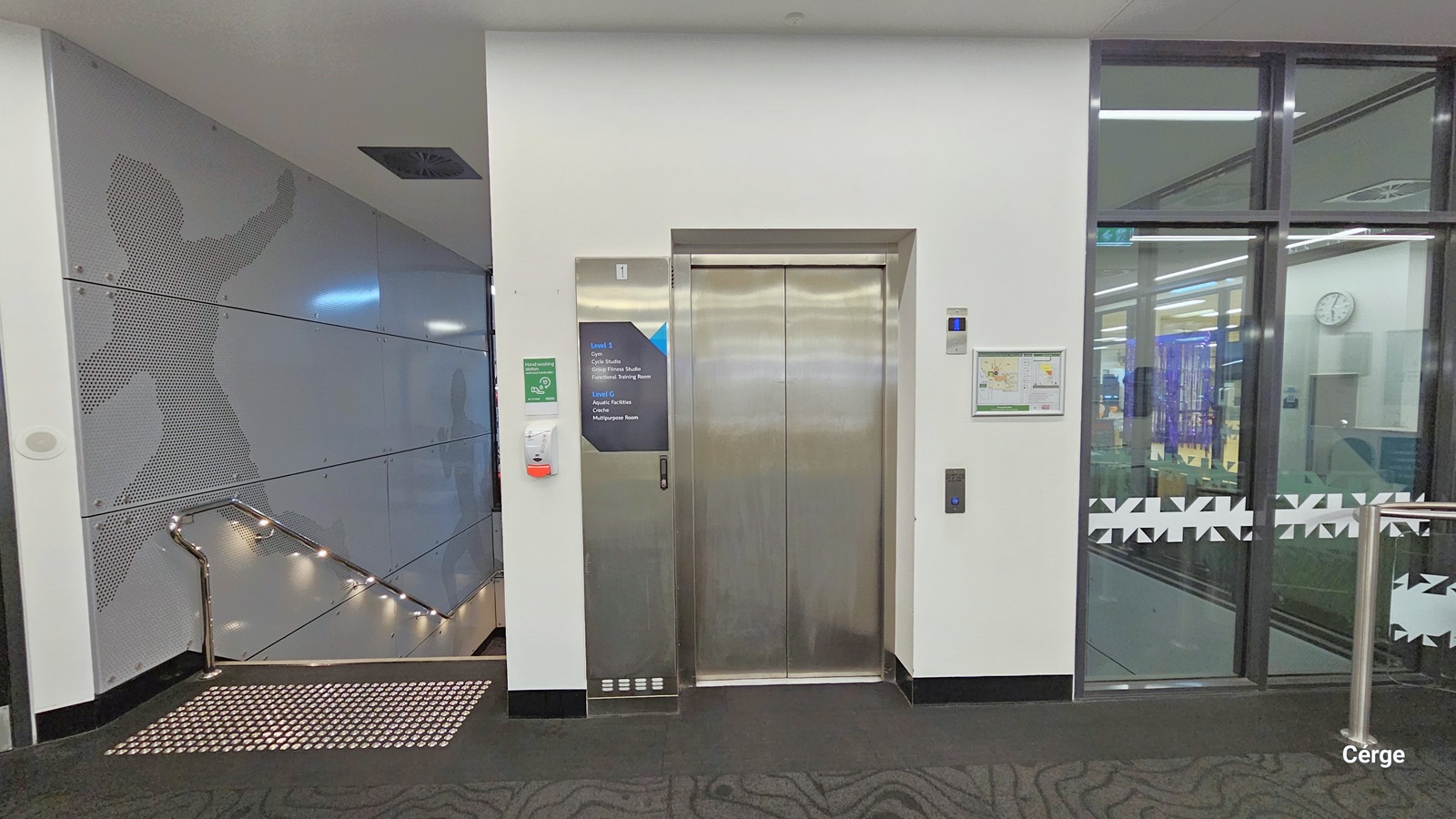 In the middle, there's a lift with wall-mounted signs indicating direction, along with a wall-mounted hand sanitizer. To the left, is the entrance to the staircase, marked by white truncated domes for safety. The stairs are equipped with stainless handrails and black carpet. The grey-painted wall features a faint shadow design of a running man. To the right, there's a glass door leading to the gym and studio, with white-painted walls and grey jigsaw print carpeting.