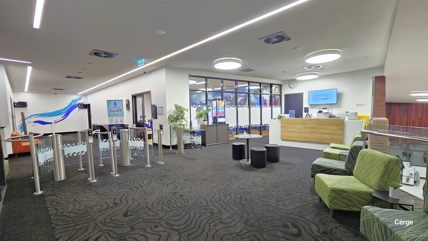 Entry to Health Club. Turnstiles are installed at the entrance. To the right is the cycle studio. There is a reception desk on the right. A round table and round cushioned stool are in the center, with cushioned green chairs lined up on the sides. The area has grey carpeted floor, an all-white ceiling, and white walls with minimal blueprints. The ceiling features designed lights and a few centralized air conditioning systems.