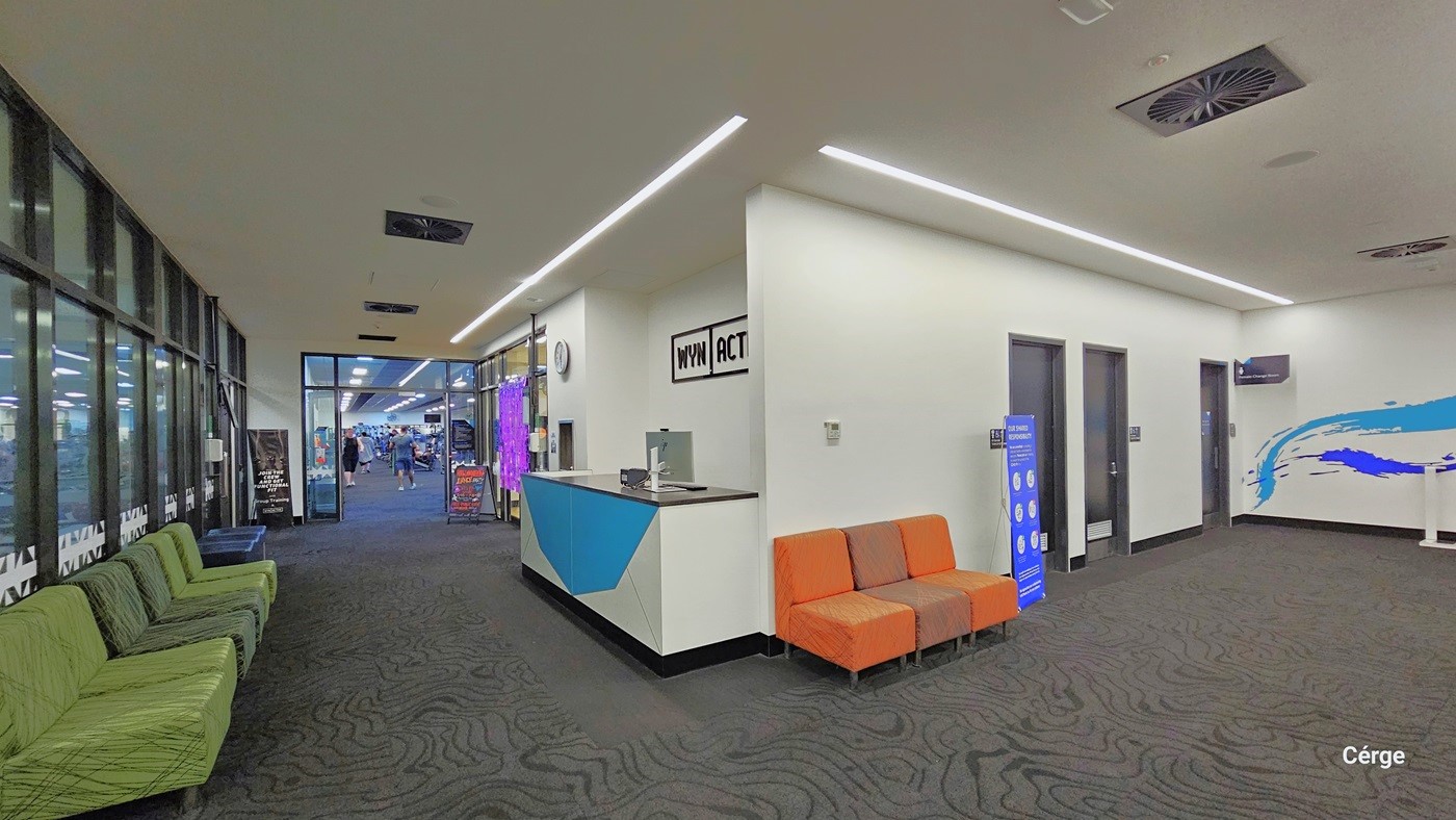To the right, there are black doors, two of which lead to all-gender toilets. In the middle, there are three cushioned orange chairs and a reception desk. On the left, a few green cushioned chairs are lined up against the glass wall. The area has grey carpeted floor, an all-white ceiling, and white walls with minimal blue prints. The ceiling features designed lights and a few centralized air conditioning systems.