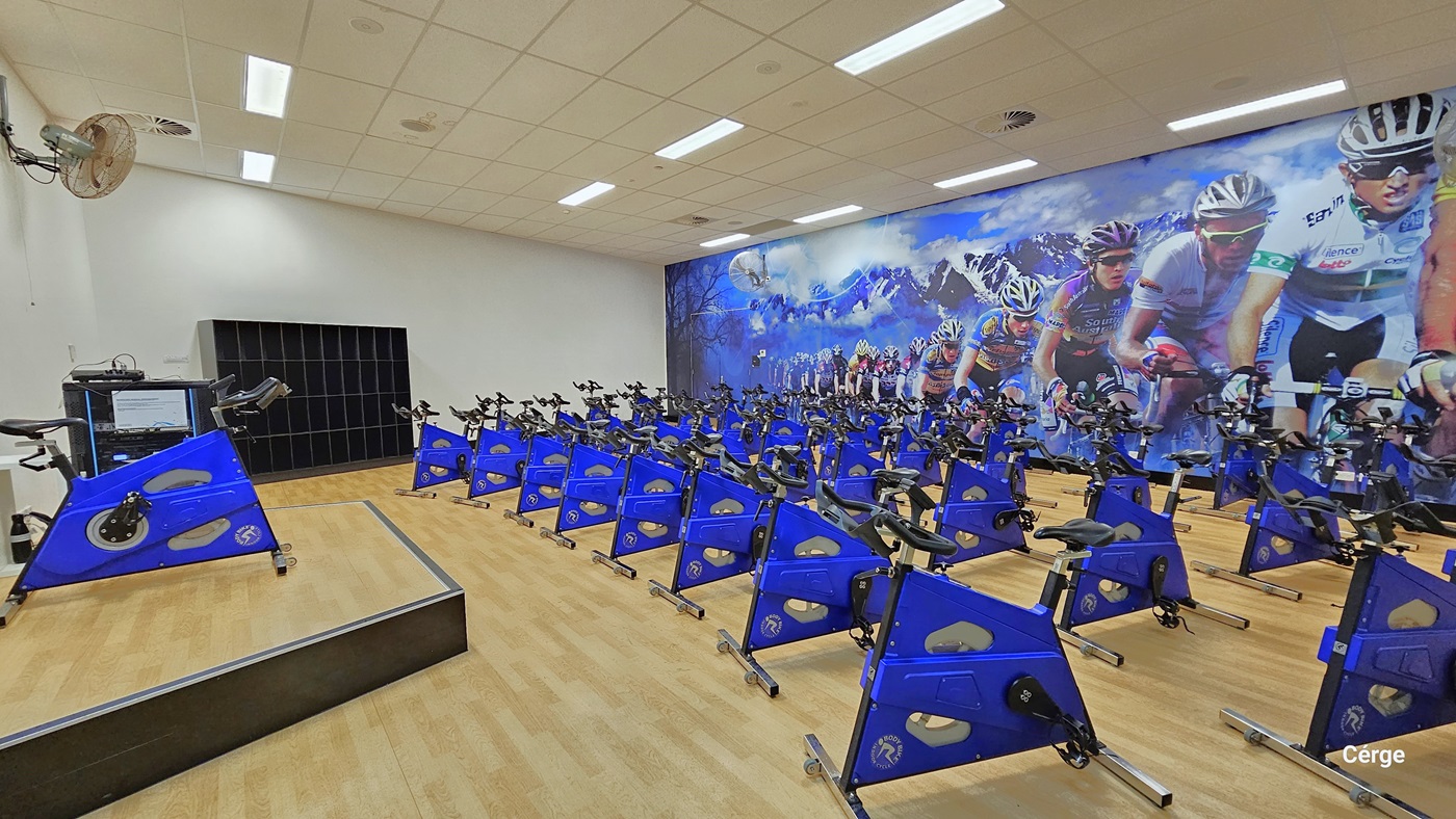 Inside the cycle studio, a row of blue cycle machines is neatly arranged, covering half of the timber floor. On the left, there is an elevated platform with a cycle machine on top, and a fan is mounted on the white wall. An open 4-tier storage cabinet is positioned against the wall. On the right, a large poster, depicting cyclists in motion, covers the entire wall. The area is well-illuminated, with white walls and a white ceiling.