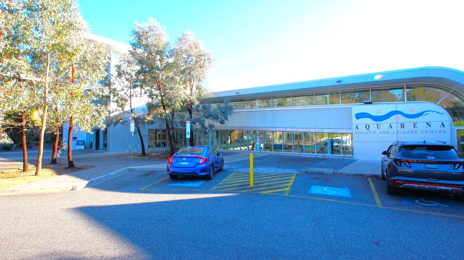 A modern building with a curved roof on the right, featuring large windows and a sign reading "Aquarena Aquatic and Leisure Centre." Two cars are parked in front, one blue and one black, with a disabled parking space marked on the ground. On the left, several tall trees line the pavement, casting shadows on the ground. The area is well-lit, and the road in the foreground is smooth and wide.