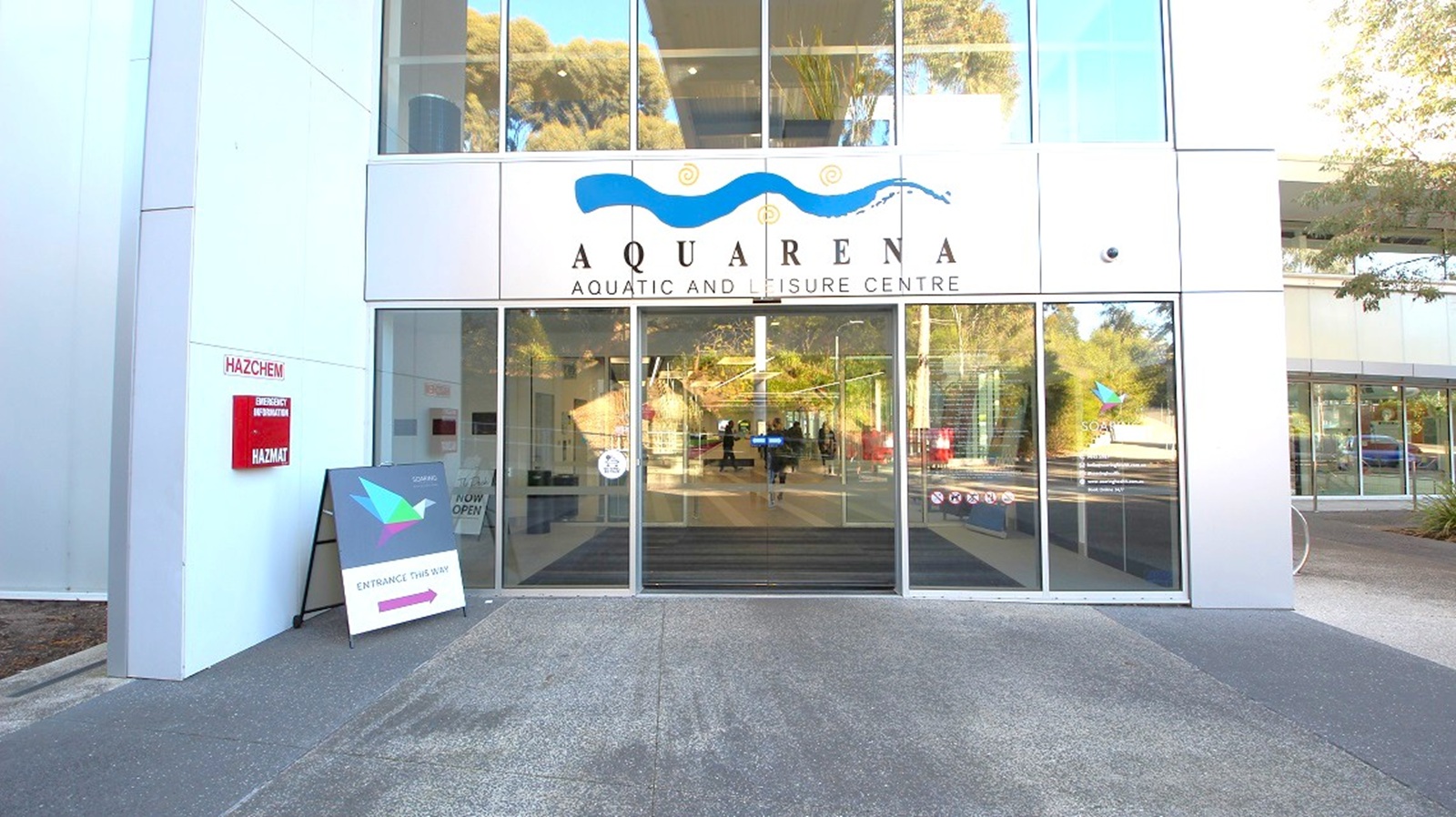 Entrance to a building with large glass doors and windows. Above the doors, a sign reads "Aquarena Aquatic and Leisure Centre" with a wave design. To the left, a red sign indicates "Hazchem" and "Mazmat," and a sandwich board sign is placed on the ground. The floor is paved, and reflections of trees and the surroundings are visible in the glass. Trees and greenery are seen in the background through the glass.