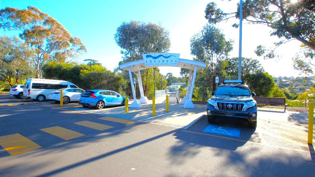 A parking area with several vehicles, including a white van and a blue car on the left, and a black SUV on the right. In the centre, a white archway with a sign reading "Quarry" leads to a pathway. Yellow bollards are in front of the archway. Trees and greenery surround the area, with a bench visible on the right. The ground is marked with yellow pedestrian lines, and a tall streetlight is on the right.