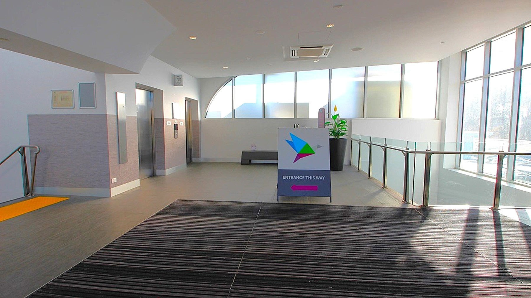 A modern indoor space with a striped carpeted floor in the foreground. On the left, there are two lift doors and a staircase with a yellow safety strip. In the centre, a sign with a colourful logo and text directs to an entrance. The right side features large windows allowing natural light to flood in, with a glass railing and a potted plant nearby. The ceiling is white with recessed lighting and an air conditioning unit.