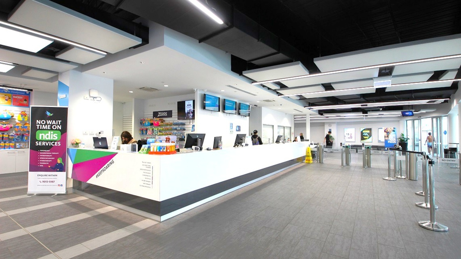 A modern interior with a long white reception desk on the right, equipped with computers and attended by staff. Above the desk are several digital screens. On the left, a colourful banner advertises "NDIS Services." The floor is grey with a tiled pattern, and the ceiling features a grid of black and white panels with integrated lighting. In the background, there are posters on the walls and a glass entrance with people entering or exiting. Queue barriers are set up in front of the desk.